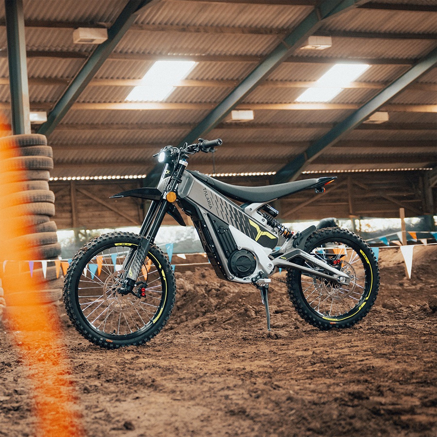 talaria x3 electric motorcycle 1907mm x 805mm x 1102mm to inches, shown on a dirt track inside an indoor riding area with banners and tires